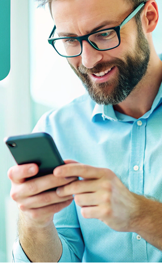 Smiling man with glasses using a smartphone, wearing a light blue shirt, in a bright indoor setting.